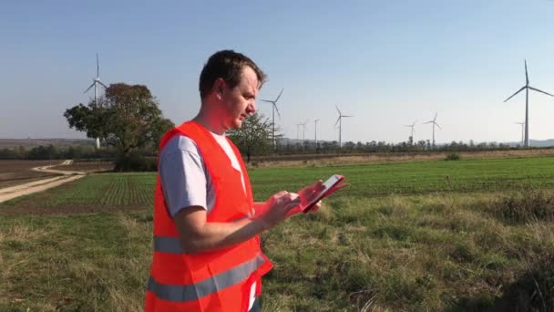 Technical engineer in safety vest making notes in tablet and looking up at wind turbine towers with rotating blades in field on windy sunny day. Technician working with touch-sensitive tablet and and entering data in project interface - Footage, Video