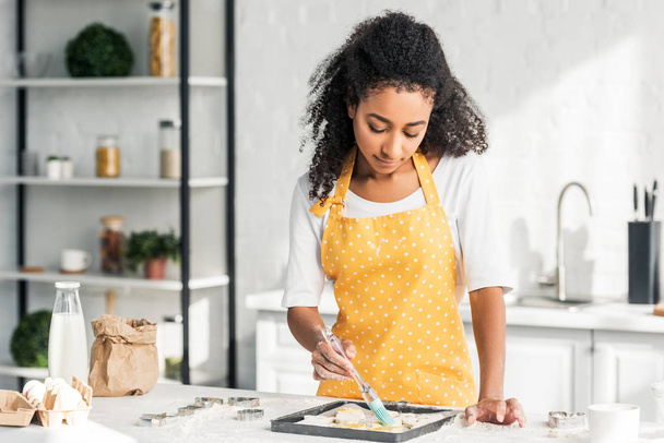 attractive african american girl in apron applying oil on unbaked cookies in kitchen - Foto, Imagen