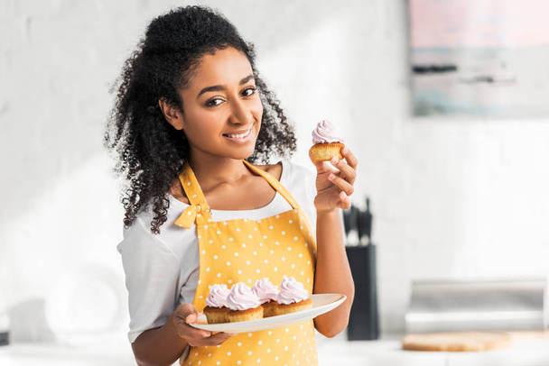 smiling attractive african american girl in apron holding homemade cupcakes and looking at camera in kitchen - Foto, afbeelding
