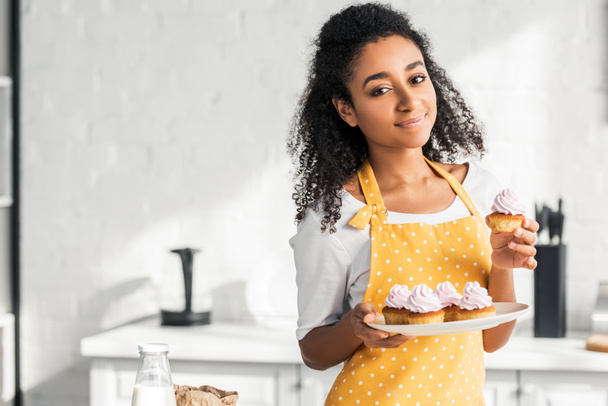 cheerful beautiful african american girl in apron holding homemade cupcakes and looking at camera in kitchen - Foto, immagini