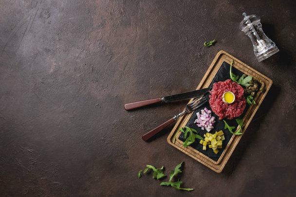 Beef tartare with quail egg in shell, cutting pickled cucumbers, capers, red onion, chives, arugula served on wooden black slate board with cutlery on dark texture background. Flat lay, space - Фото, зображення