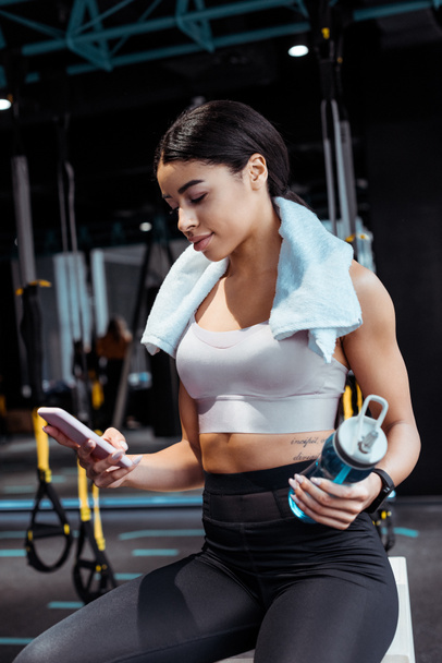Beautiful sportive girl using digital device while holding sport bottle in gym  - Photo, Image