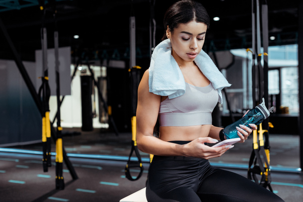 Menina esportiva atraente usando smartphone enquanto segurando garrafa de esporte no ginásio esportivo
  - Foto, Imagem