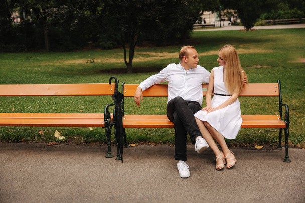 schönes Paar sitzt auf der Bank im Park. Blonde Haare Mann und Frau verbringen Zeit miteinander. Sie nehmen einen schwarzen Regenschirm. Hintergrund des grünen Grases - Foto, Bild