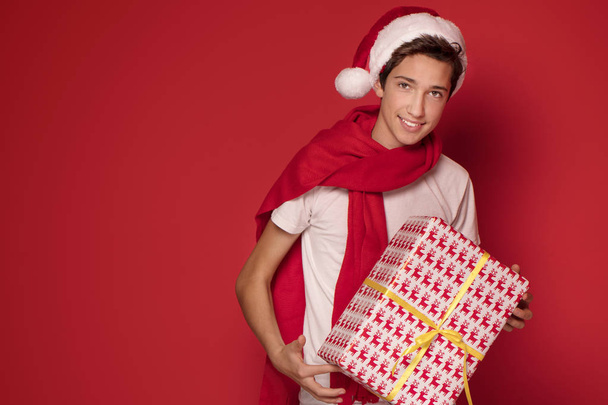 Happy teenager boy with Christmas gift posing in Santa Claus hat, smiling, red studio background. - Фото, зображення