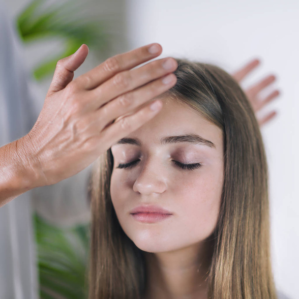 Afbeelding van de horizontale van vrouwelijke Reiki therapeut permanent en hand in hand boven het hoofd van de mooie tiener patiënt. Reiki Practitioner transfering energie en Helen kruinchakra. Vreedzame meisje zit met haar ogen dicht - Foto, afbeelding