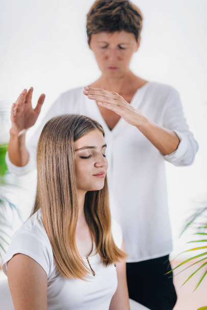 Vaakasuora kuva naispuolisesta Reiki-terapeutista seisomassa ja pitämässä kädestä kauniin teini-ikäisen potilaan pään yläpuolella. Reiki Practitioner siirtää energiaa ja parantavaa kruunu chakra. Rauhallinen tyttö istuu silmät kiinni
 - Valokuva, kuva