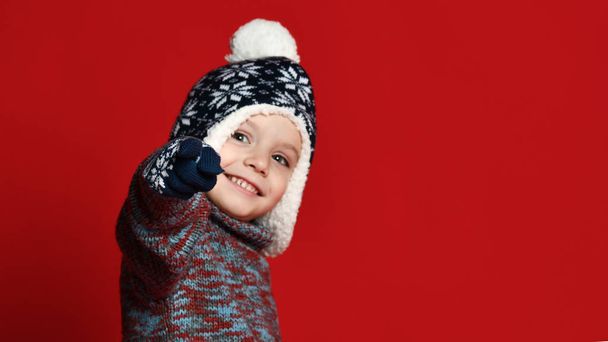 child boy in knitted hat and sweater and mittens having fun over colorful red  background. - Photo, image