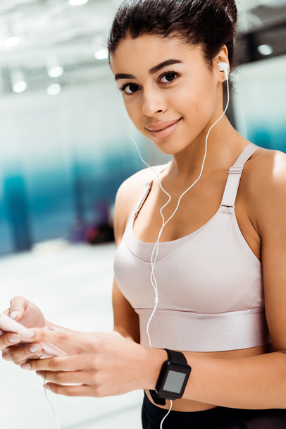 Attractive sportive girl smiling at camera while listening to music in fitness gym - Foto, Imagem