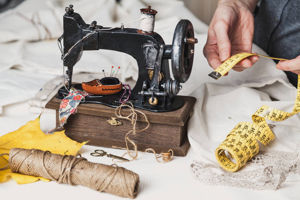 Workplace seamstress. Vintage sewing machine and a skein of threads. A yellow maple leaf lies next to it. The woman's hands fold a flexible tap - Φωτογραφία, εικόνα