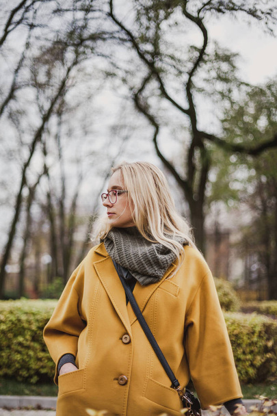 beautiful blonde in glasses and walking in an autumn park on her bright yellow fashion coa - Foto, afbeelding