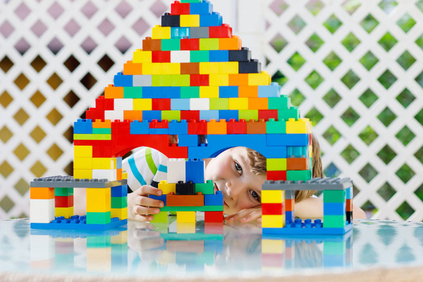 Egy kis szőke gyerek, aki sok színes műanyag tömbbel játszik. Imádnivaló óvodás fiú visel színes ing és szórakozás épület nagy kastély és hozzon létre egy házat - Fotó, kép