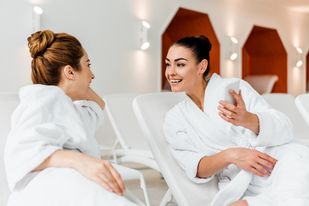 happy young woman showing smartphone to girlfriend while resting together on sunbeds in spa - Photo, image