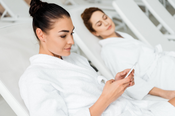 smiling young woman in bathrobe using smartphone while resting on sunbed in spa  - Photo, image