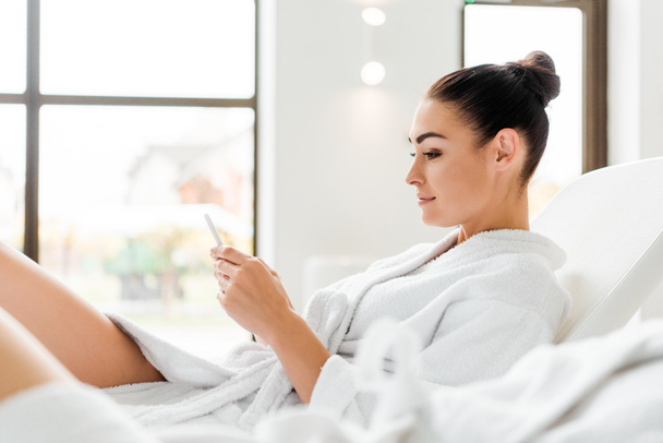 side view of smiling young woman in bathrobe using smartphone while resting in spa - Photo, Image