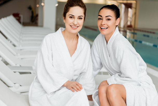 happy young women in bathrobes smiling at camera while relaxing together in spa - Foto, afbeelding