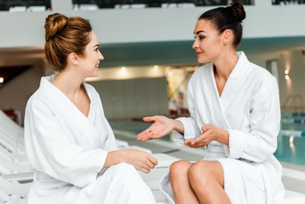 smiling young women in bathrobes talking while relaxing together in spa - Foto, afbeelding