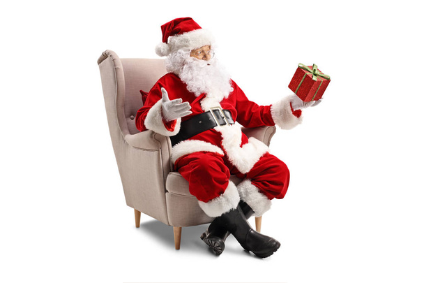 Kerstman zitten in een leunstoel en een cadeautje geïsoleerd op een witte achtergrond te houden - Foto, afbeelding