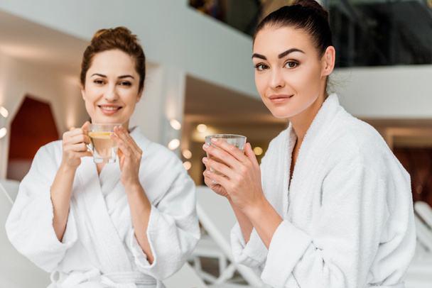beautiful young women in bathrobes holding cups with herbal tea and smiling at camera in spa - 写真・画像