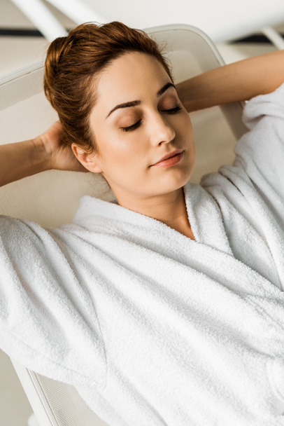 attractive young woman in bathrobe lying with hands behind head and closed eyes in spa  - Foto, Imagem