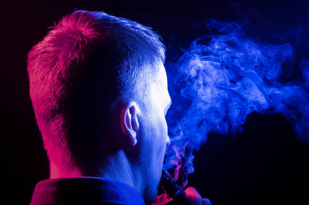 Вид ззаду голови чоловіка в сорочці, що курить вейп і видихає різнокольоровий дим синього кольору з правого боку від себе на чорному ізольованому тлі. Шкода здоров'ю людини
 - Фото, зображення