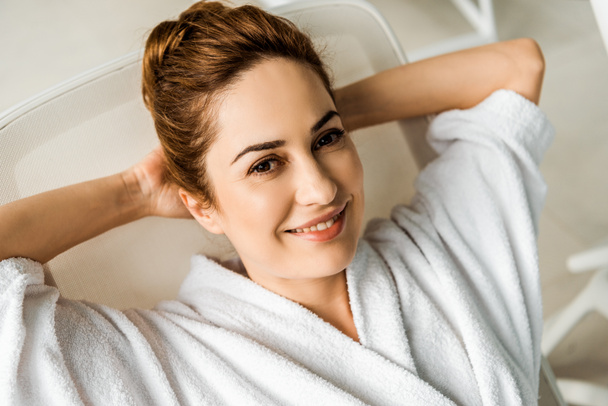 heureuse jeune femme en peignoir couché avec les mains derrière la tête et souriant à la caméra dans le spa
  - Photo, image