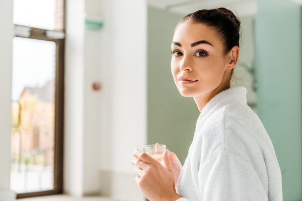 attraktive junge Frau im Bademantel mit einer Tasse Kräutergetränk und Blick in die Kamera im Wellnessbereich - Foto, Bild