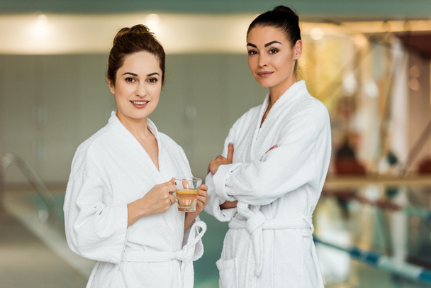 beautiful women in bathrobes holding cup with herbal tea and smiling at camera in spa - Fotó, kép