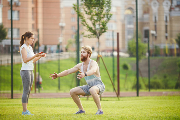 Spor takım çalışması. kadın eğitmen Eğitim Parkı yeşil çimenlerin üzerinde çim batımında sakallı adam. Kız telefon elinde tutan ve kronometre kullanır, adam oturarak kalça için egzersizler - Fotoğraf, Görsel