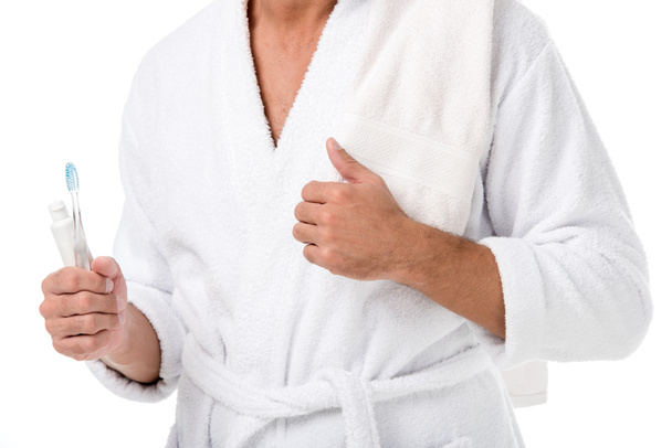 περικομμένη εικόνα του ανθρώπου στο μπουρνούζι με πετσέτα πάνω από τον ώμο κρατώντας οδοντόκρεμα και οδοντόβουρτσα που απομονώνονται σε λευκό - Φωτογραφία, εικόνα