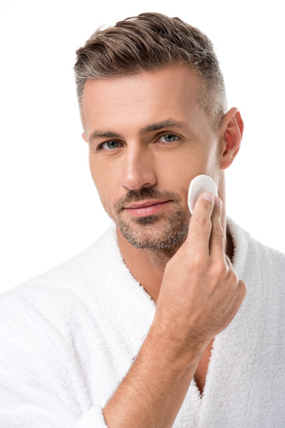 portrait of handsome man in bathrobe applying tonic by cotton pad on face isolated on white - Photo, Image
