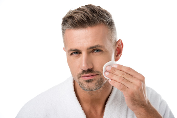 portrait of handsome man in bathrobe applying tonic by cotton pad on face isolated on white - Foto, immagini