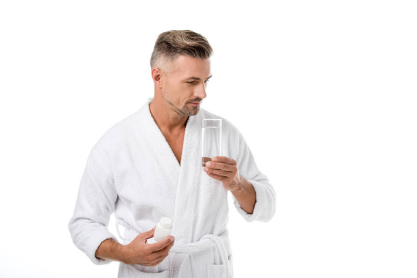 örömteli felnőtt ember fürdőköpeny mutatja a vitaminok és a gazdaság pohár vizet elszigetelt fehér - Fotó, kép