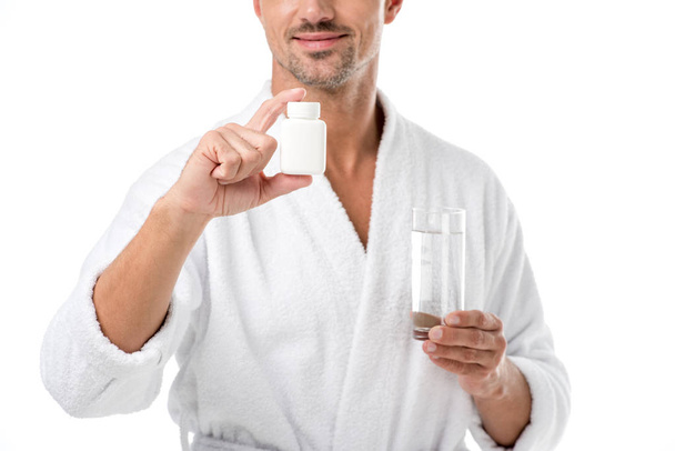 částečný pohled dospělého člověka v županu ukazující vitamíny a držení sklenice vody izolované na bílém - Fotografie, Obrázek