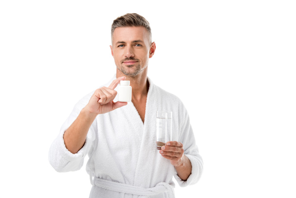 joyful adult man in bathrobe showing vitamins and holding glass of water isolated on white - Photo, Image