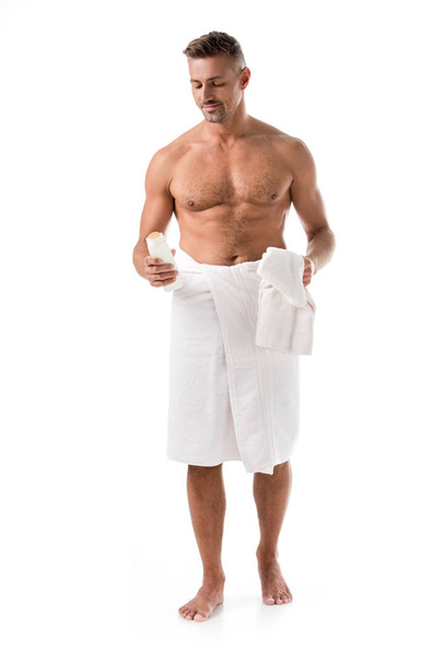 fröhliche muskulöse hemdlose Mann in Handtuch eingewickelt hält Duschgel isoliert auf weiß - Foto, Bild