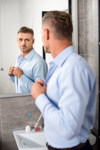 rear view of adult businessman looking at mirror while buttoning up blue shirt in bathroom at home - 写真・画像