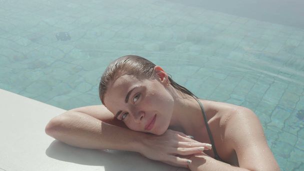 Молода красива жінка дивиться в камеру, розслабляючись на краю відкритого басейну
 - Фото, зображення