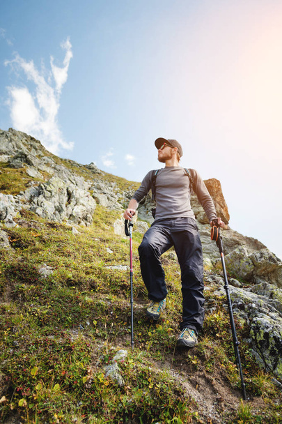 A bearded man in sunglasses and a cap with a backpack stands on top of a rock and looks into a rocky valley high in the mountains. The concept of tourism and easy trekking in the mountains outdoor - Valokuva, kuva