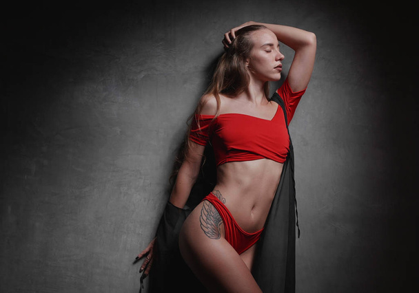 Beautiful young woman in red bikini and black overall posing over gray wall background - 写真・画像