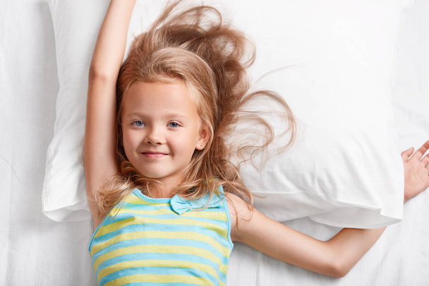 Above view of attractive light haired small child, has blue eyes, stretches in bed, lies on white pillow, dressed in colourful striped pyjamas, enjoys recreation time in bedroom. Children and rest - Photo, image