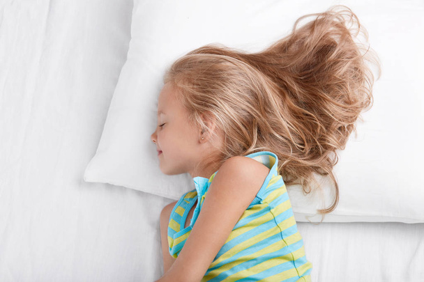 Sideways shot of small kid wears nightwear, being deep in sleep, rests at bed, lies on white bed, enjoys bedtime, rests after active games outdoor with friends, naps in bedroom. Children concept - Fotografie, Obrázek