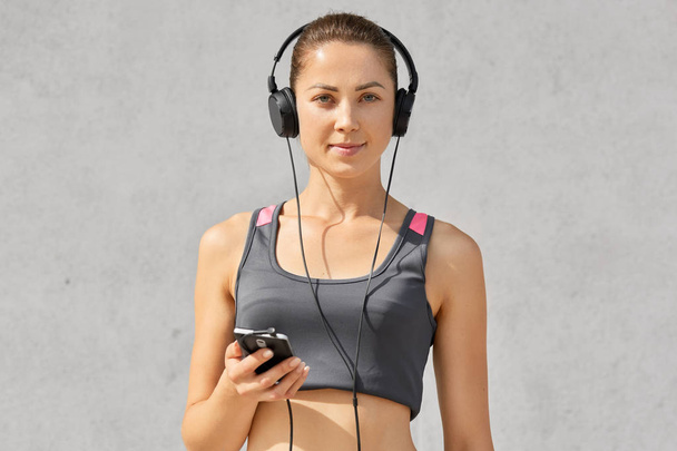 Isolated shot of young sportswoman has healthy athleteic body, listens music with headphones, holds smart phone, looks directly at camera, has cardio training, poses against grey concrete wall - Photo, image