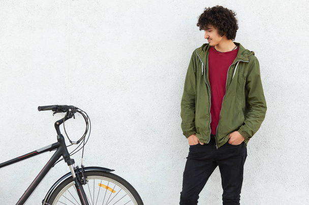 Photo of attractive hipster with curly dark hair, wears green jacket and black trousers, keeps hands in pocket, looks at his bike after ride, isolated over white concrete background. Youth concept - Φωτογραφία, εικόνα
