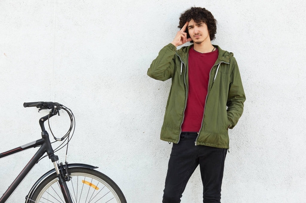 Pensive man keeps finger on temples, tries to recollect something in mind, dressed in green jacket, stands near bicycle, isolated over white concrete background. People, youth and transport concept - Foto, imagen