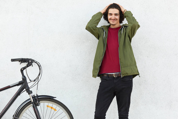 Photo of handsome content unshaven guy looks with positive expression at camera, keeps hands on head, wears fashionable jacket, stands near bicycle indoor. People, lifestyle and fashion concept - Foto, Imagen