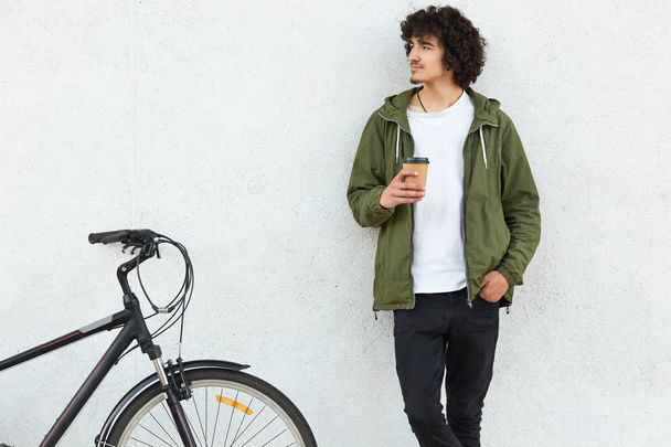 Photo of handsome youngster with curly bushy hair, dressed in white t shirt, trousers, green anorak, keeps hand in pocket, concentrated aside, drinks takeaway coffee, rides on sport bicycle. - Valokuva, kuva