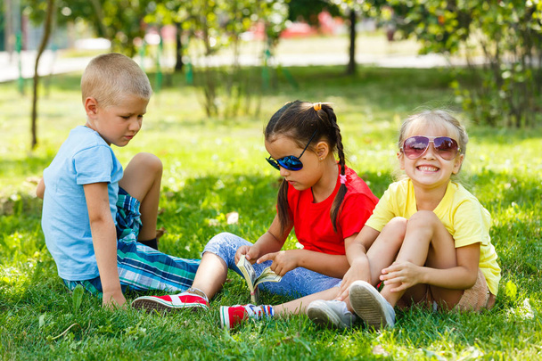 Δύο μικρά κορίτσια και ένα αγόρι με μια στοίβα από δολάρια που κάθονται σε πράσινο γρασίδι στο πάρκο - Φωτογραφία, εικόνα