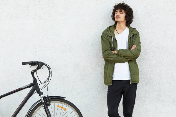 Self confident focused man has curly hairstyle, keeps arms folded, dressed in jacket and jeans, looks thoughtfully aside, rides bicycle, models against white background. Active lifestyle concept - 写真・画像
