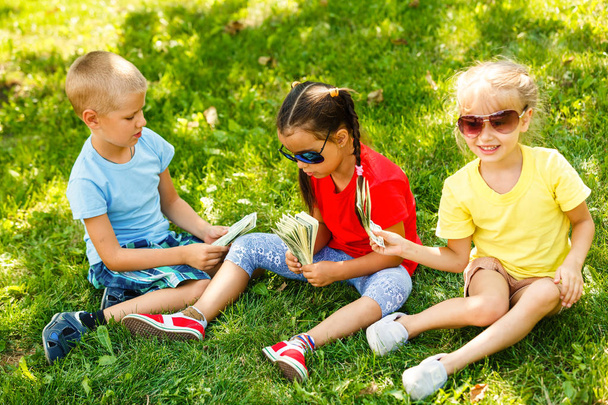 公園の緑の草の中に座ってドルの山を持つ2人の小さな女の子と少年 - 写真・画像